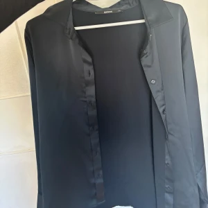 Svart skjorta från BikBok - Stilren svart skjorta från BikBok med klassisk krage och knäppning framtill. Endast använt en gång. Skjortan har långa ärmar och är tillverkad i ett slätt, lätt glansigt material som ger en dressad känsla. 