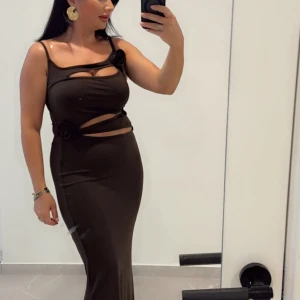 Lång brun klänning med cut-outs och blommor - Säljer en lång, mörkbrun klänning med smala axelband och unika cut-outs vid midjan och bröstet. Klänningen har dekorativa blommor som detaljer och en tight passform som framhäver figuren.