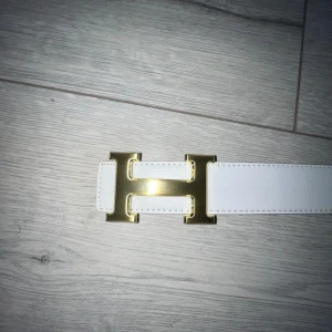 Vitt skärp med guldspänne från  - Säljer ett stilrent vitt bälte från  med ett stort -format spänne i guld. Bältet är gjort i skinn och har en klassisk, rak form. Perfekt accessoar för att lyfta din outfit.