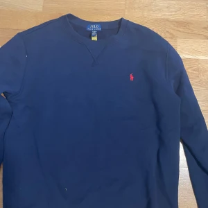 Mörkblå sweatshirt från Polo Ralph Lauren - Mörkblå sweatshirt från Polo Ralph Lauren med klassisk rund halsringning och röd broderad logga på bröstet. Tröjan har långa ärmar och är tillverkad i mjukt material, perfekt för en stilren och avslappnad look.