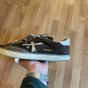 Bruna sneakers från Premiata - Säljer ett par bruna sneakers från Premiata med beige detaljer och vit sula. Skorna har snörning och är tillverkade i mocka med märkets logga på sidan. Perfekta för en stilren vardagslook.