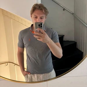 Grå pikétröja från Polo Ralph Lauren - Säljer en klassisk grå pikétröja från Polo Ralph Lauren med orange detaljer på insidan av kragen och broderad logga på bröstet. Perfekt för en stilren och avslappnad look. 
