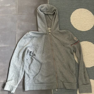 Grå hoodie med dragkedja från Moncler - Snygg grå hoodie från Moncler med dragkedja framtill och klassisk logga på ärmen. Tröjan har huva, fickor och en diskret randdetalj i rött, vitt och blått på ärmen. Perfekt för en avslappnad och stilren look.