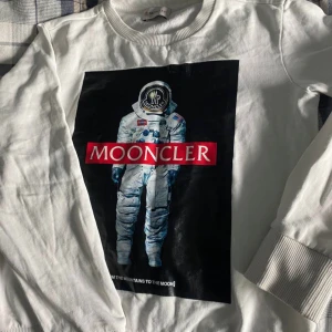 Vit sweatshirt med astronaut-tryck från Moncler - Säljer en vit sweatshirt från Moncler med ett stort tryck av en astronaut och texten 'MOONCLER' på bröstet. Tröjan har långa ärmar och ribbade muddar vid ärmslut och nederkant. Perfekt för dig som gillar unika prints och streetwear. Pris ej hugget i sten