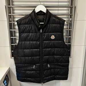 Moncler Gui Storlek 3 - Snygg svart dunväst från Moncler med hög krage och två framfickor med dragkedja. Västen har Moncler-logga på bröstet och stängs med dragkedja framtill. Perfekt för lager-på-lager och stilren look. Västen är sparsamt använd och köpt på monclers hemsida. Originalboxen med alla tillbehör ingår i köpet. Skriv om ni har frågor.