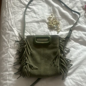 Maje väska  - Snygg olivgrön maje väska i mocka med dekorativa fransar på sidorna. Väskan har ett stilrent handtag upptill och en lång axelrem. Perfekt för att addera lite edge till din outfit.