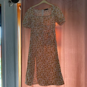 Blommig sommarklänning med puffärm - Söt vit klänning med gula och blå blommor, fyrkantig ringning och kort puffärm. Klänningen har slits framtill och är i midi-längd, perfekt för varma dagar.