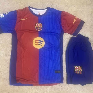 FC Barcelona matchtröja Lamine Yamal #19 - Säljer en officiell FC Barcelona fotbollströja med Lamine Yamal och nummer 19 på ryggen. Tröjan är kortärmad, har röd och blå färg med klubbmärke och sponsorlogga på bröstet. Ingår matchande blå shorts med klubbmärke.