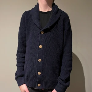 Mörkblå kofta från Cocodrillo - Säljer en snygg mörkblå kofta från Cocodrillo med stora träknappar. Koftan är stickad och har en skön krage som ger extra värme. Perfekt för kyliga dagar. Passar bra till både jeans och chinos.