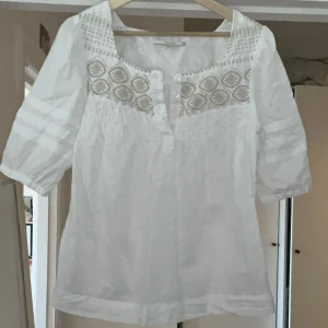 Vit blus med broderade detaljer från Part Two - Säljer en luftig vit blus från Part Two med broderade mönster upptill och halvknäppning framtill. Blusen har trekvartsärmar med dekorativa ränder och en lös passform. Perfekt för vår och sommar!