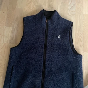 Mörkblå fleeceväst med dragkedja - Mysig mörkblå fleeceväst med hög krage och hel dragkedja framtill. Västen har en liten broderad logotyp på bröstet och är perfekt för lager-på-lager. Passar både till promenader och chill.