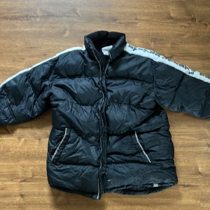 Svart pufferjacka med vita detaljer - Säljer en svart pufferjacka från Kappahl med vita ränder och text längs ärmarna. Jackan har dragkedja framtill, höga krage och två fickor med dragkedja. Perfekt för kyligare dagar och riktigt skön att bära.