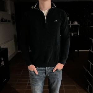 Svart halvzip-tröja från GANT - Säljer en svart långärmad halvzip-tröja från GANT med vit rand vid kragen och diskret logga på bröstet. Tröjan har en klassisk passform och är tillverkad i ett mjukt bomullsmaterial. Perfekt för en stilren och avslappnad look.