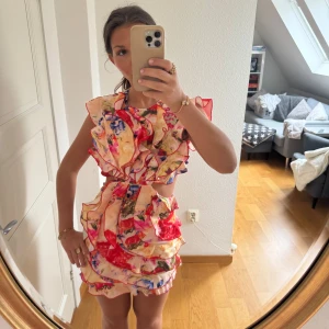 Färgglad volangklänning med blommigt mönster - Säljer en kort, ärmlös klänning med volanger och blommigt mönster i rött, blått, gult och rosa. Klänningen har öppen rygg och markerad midja. Perfekt för dig som vill sticka ut med färg och volym! Materialet känns lätt och luftigt.