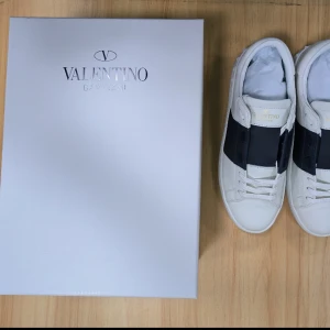 Valentino Garavani vita sneakers med svart detalj - Säljer ett par vita Valentino Garavani sneakers med bred svart rem över mitten. Klassisk design med rund tå och platt sula. Perfekta för dig som gillar exklusiva och stilrena skor.