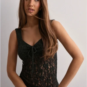 Svart spetslinne med knyt från NELLY  - Säljer ett svart, ärmlöst spetslinne med v-ringning och knyt framtill. Toppen har ett genombrutet spetsmönster som ger en snygg och trendig look. Perfekt att styla till olika outfits. Aldrig använd! Mindre i storlek