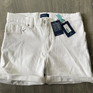 Vita jeansshorts från GANT - Snygga vita jeansshorts från GANT med klassisk femficksdesign och uppvikta benslut. Perfekta för sommaren och lätta att matcha med olika toppar. Stängs med knapp och dragkedja framtill. Prislappen sitter kvar, då de aldrig har kommit till användning!