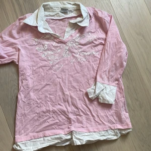 Ljusrosa långärmad tröja med vit krage från Comfort Fashion - Säljer en ljusrosa långärmad tröja med vit skjortkrage och vita manschetter från Comfort Fashion. Tröjan har ett broderat mönster och text framtill. Perfekt för dig som gillar en preppy stil med extra detaljer.