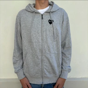Cdg hoddie  - Grå hoodie med dragkedja från Comme des Garçons Play. Tröjan har ett svart hjärta med ögon på bröstet och ett större svart hjärta med ögon på ryggen. Utrustad med huva och fickor framtill. Perfekt för en avslappnad stil. Den är storlek L men passar M. 