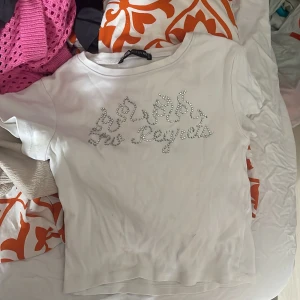 Vit t-shirt med strasstext från B Sister - Vit t-shirt från B Sister med korta ärmar och glittrig strasstext framtill. T-shirten har en klassisk passform och är tillverkad i mjukt material, perfekt för en trendig vardagslook.