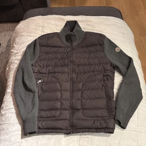 Grå jacka från Moncler - Snygg grå jacka från Moncler med quiltad front och stickade ärmar. Jackan har en dragkedja framtill och ribbade muddar. Perfekt för kyligare dagar.