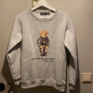 Grå sweatshirt med Polo Bear från Ralph Lauren - Säljer en grå sweatshirt från Ralph Lauren med det ikoniska Polo Bear-motivet på framsidan. Tröjan har långa ärmar och en rund halsringning. Perfekt för en avslappnad stil med en touch av lyx.