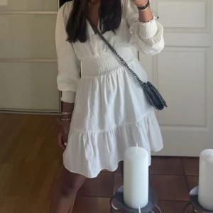 Vit klänning med v-ringning - Säljer en söt vit klänning med v-ringning och långa ärmar. Klänningen har en smockad midja och volangdetaljer vid fållen. Fint skick! Pris kan diskuteras 