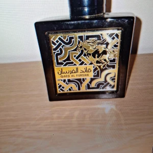 Qaed Al Fursan Parfym - Elegant parfymflaska i svart med gyllene detaljer och ett stiliserat hästmotiv. Flaskan har en fyrkantig form och en svart kork. Perfekt för den som söker en unik och kraftfull doftupplevelse.