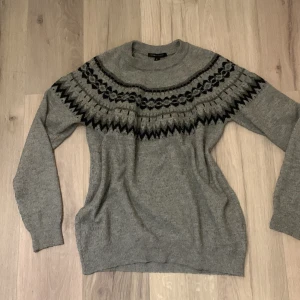 Grå stickad tröja från MyWearWoman - Säljer en mysig grå stickad tröja från MyWearWoman med ett snyggt mönster i svart och mörkgrått runt halsen. Tröjan har långa ärmar och en ribbad krage. Perfekt för kyliga dagar!