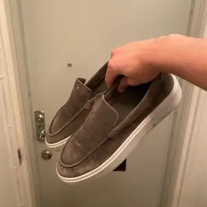 Säljer ett par grön-bruna loafers med vit sula. Skorna har en stilren design utan snörning, vilket gör dem enkla att ta på och av. Perfekta för en avslappnad stil.