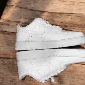 Vita Nike Air Force sneakers - Säljer ett par klassiska vita Nike Air Force sneakers. De har en stilren design med snörning och den ikoniska Nike-loggan på sidan och hälen. Perfekta för en casual look.