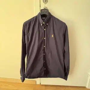Snygg mörkblå skjorta från Polo Ralph Lauren med klassisk krage och knappar framtill. Den har ett broderat gult märke på bröstet och långa ärmar med knappar vid ärmsluten. Perfekt för en stilren look!