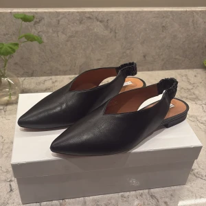 Svarta slip-ins från & Other Stories - Stilrena svarta slip-ins från & Other Stories i skinn med spetsig tå och elastisk hälrem. Perfekta för en chic och bekväm look. Använda 1 gång💕
