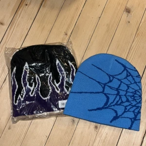 Två stickade mössor med mönster - Säljer två coola mössor. Den ena är svart med vita och lila flammor, och den andra är blå med ett spindelnätsmönster. Perfekta för att ge din outfit en unik touch!