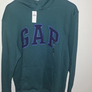 Grön hoodie från GAP - Säljer en snygg grön hoodie från GAP med stor logga på bröstet. Perfekt för en avslappnad stil. Den har en klassisk känguruficka och en bekväm huva.