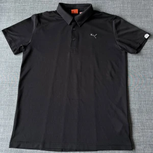 Svart pikétröja från Puma - 📦Säljer en stilren svart pikétröja från Puma med klassisk krage och knappar framtill. Tröjan har korta ärmar och ett diskret Puma-logotyp på bröstet. Perfekt för en sportig look eller avslappnad stil. 📐(Storlek M, normal i storleken) Inga större defekter än det som syns på bilden, något använd men sparsamt. Vid frågor, tveka inte att höra av er! ☺️