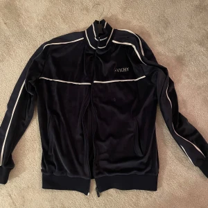 Svart träningsjacka från Vichy - Snygg svart träningsjacka från Vichy med vita detaljer. Jackan har dragkedja och långa ärmar, perfekt för en sportig look. Den har en stilren design med ribbade muddar vid ärmslut och krage.