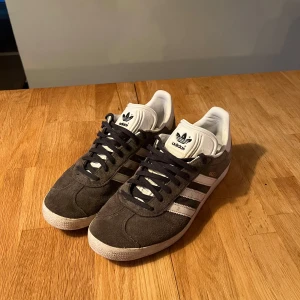 Adidas Gazelle i grå mocka - Säljer ett par klassiska Adidas Gazelle sneakers i grå mocka med vita detaljer. Har haft skorna en sommar så är i ett 8/10 skick.                                      Nypris: 1200 storlek 40 passar 41