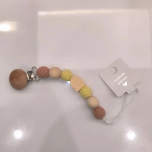 Napphållare med träkulor - Charmig handgjord napphållare med träkulor i beige, brun och gul. Den har en praktisk klämma i metall och en söt regnbågsdetalj i mitten. Perfekt för att hålla nappen nära till hands. Kan göra med namn vid beställning.