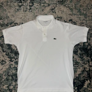 Vit pikétröja från Lacoste - Säljer en klassisk vit pikétröja från Lacoste med den ikoniska krokodilloggan på bröstet. Tröjan har korta ärmar och en knappslå framtill. Perfekt för en stilren och avslappnad look.