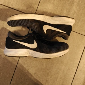 Svarta Nike sneakers - Säljer ett par svarta Nike sneakers med vit sula och den klassiska swoosh-loggan i vitt. Skorna har snörning och är perfekta för träning eller vardagsbruk.