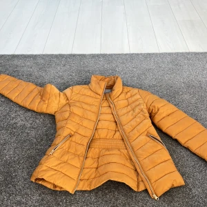 Orange quiltad jacka från Lindex - Säljer en snygg orange quiltad jacka från Lindex. Jackan har en dragkedja framtill och två praktiska fickor med dragkedjor. Perfekt för höst och vår med sin lätta och bekväma design.
