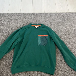 Grön tröja från H&M Move - Säljer en grön tröja från H&M Move med en unik bröstficka i en annan grön nyans och en orange kant. Tröjan har långa ärmar och en bekväm passform. Perfekt för en avslappnad stil.
