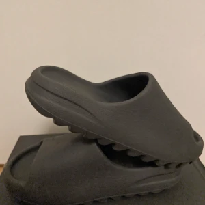 Yeezy slide onyx från adidas - Snygga svarta sandaler med en enkel och stilren design. De har en tjock sula med räfflad undersida för extra grepp. Perfekta för en avslappnad stil. Perfekt nu inför sommaren. De ser i princip helt nya ut men de är använda en sommar.