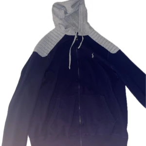 Ralph lauren zip - Snygg svart hoodie med grå huva och axelparti. Den har en dragkedja framtill och justerbara snören vid huvan. Perfekt för en avslappnad stil. Ny pris 1800 