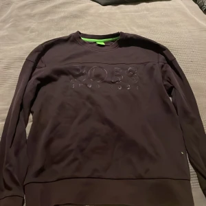 Svart tröja från Hugo Boss - Säljer en stilren svart tröja från Hugo Boss med deras logga i relief på framsidan. Tröjan har långa ärmar och en rund halsringning med en grön detalj på insidan av kragen. Perfekt för en casual look. 