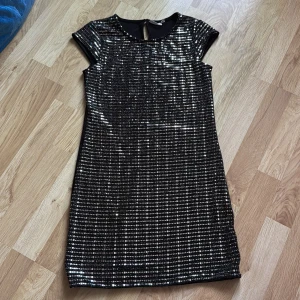 Svart glittrig klänning från KappAhl - Snygg svart klänning från KappAhl med glittriga paljetter och korta ärmar. Perfekt för en festlig kväll! Klänningen har en smickrande passform och en liten knapp i nacken för enkel påklädning.