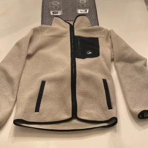 Beige fleecejacka med dragkedja - Säljer en mysig beige fleecejacka med dragkedja och svarta detaljer. Jackan har en bröstficka med dragkedja och två sidofickor. Perfekt för kyliga dagar!