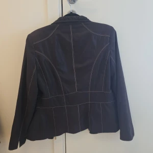 Brun skinnjacka från Kirsten - Snygg Brun  skinnjacka från Kirsten med dragkedja framtill och två fickor med dragkedjor. Jackan har en stilren design med vita sömmar som ger en cool kontrast. Perfekt för att ge din outfit en edgy touch.