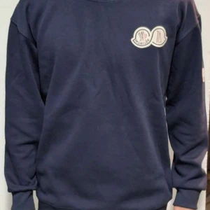 Mörkblå sweatshirt från Moncler - Säljer en stilren mörkblå sweatshirt från Moncler med deras ikoniska logga på bröstet och ärmen. Tröjan har långa ärmar och en klassisk rund halsringning. Perfekt för en avslappnad och trendig look.